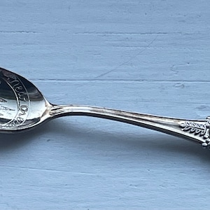 May include: Silver souvenir spoon featuring an engraved design of New Orleans landmarks on the bowl and a blue enamel shield with the text "New Orleans, LA" and a depiction of a person riding a horse on the handle.