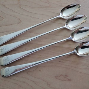 Puede incluir: Cuatro cucharas de plata con un diseño de monograma. Las cucharas tienen un mango largo y delgado y un cuenco redondeado.