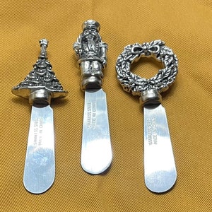 May include: Three silver Christmas-themed butter knives with detailed handles. The handles are shaped like a Christmas tree, a nutcracker, and a wreath. The blades are stainless steel and have the text "Stainless Steel Made in China" on the back.
