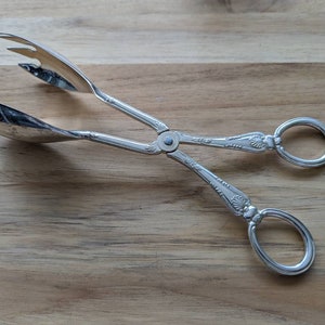 Op de afbeelding: Zilveren saladetang met een sierlijk ontwerp en twee handvatten. De tang heeft aan het ene uiteinde een lepelvorm en aan het andere uiteinde een gekartelde vorm.