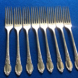 May include: Set of ten silver forks with ornate handles. The forks have a polished finish and are arranged in a row on a blue surface.