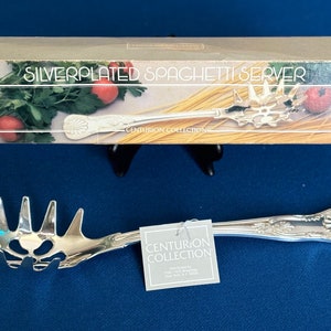 Puede incluir: Un servidor de espaguetis plateado en su caja original. La caja presenta una foto del servidor con espaguetis y una guarnición verde de hojas. La caja también dice "Silverplated Spaghetti Server" y "Centurion Collection".
