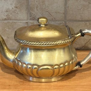 May include: A gold teapot with a hinged lid and a silver handle. The teapot has a decorative design on the body and a rope-like trim around the lid.
