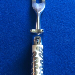 May include: A silver serving fork with a white handle featuring a silver swirl pattern. The fork has two prongs.