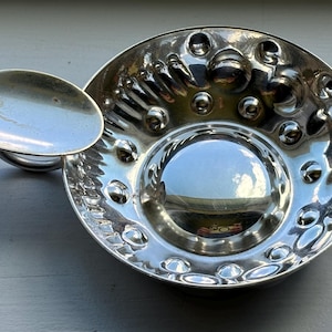 May include: A silver serving dish with a hammered texture and a small spoon attached to the side. The dish is round and has a reflective surface.
