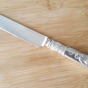 May include: A silver butter knife with an ornate handle featuring a detailed design. The handle is decorated with a raised pattern and a letter 'D'.