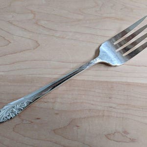 Puede incluir: Un tenedor de plata con un diseño floral adornado en el mango. El tenedor tiene cuatro dientes y está hecho de metal pulido.