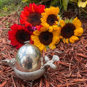 May include: A silver plated bird-shaped serving dish with a domed lid. The dish is surrounded by a bouquet of yellow, red, and orange sunflowers.