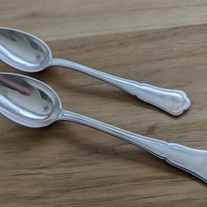 May include: Two silver spoons with ornate handles. The spoons are lying on a wooden surface.