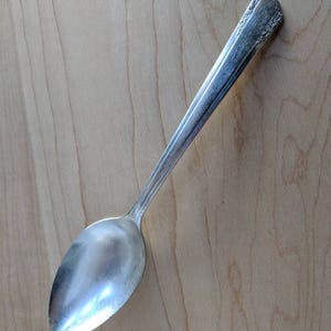 May include: A silver spoon with a rounded bowl and a long handle. The handle has a decorative pattern near the top. The spoon is set against a light brown wooden surface.