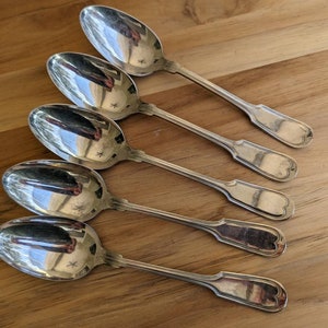 Puede incluir: Cinco cucharas de plata con mangos adornados. Las cucharas están dispuestas en fila, con los mangos apuntando hacia la derecha. Las cucharas reflejan la luz, creando un efecto brillante.