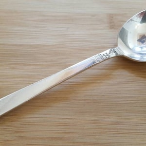 May include: A silver soup spoon with a decorative pattern on the handle. The spoon is on a wooden surface.