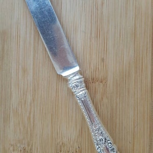 Puede incluir: Un cuchillo de mantequilla de plata con un mango adornado. El mango está decorado con un patrón floral.