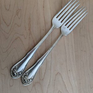 May include: Two silver forks with ornate rose designs on the handles. The forks are laid out on a light brown wooden surface.