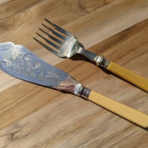 May include: A set of two vintage silver-plated serving utensils with yellow plastic handles. The knife has an ornate floral design and the fork has four tines.