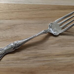 Könnte beinhalten: Eine silberne Gabel mit einem verzierten Griff mit einem Traubenmuster. Die Gabel hat vier Zinken und ist in gutem Zustand.