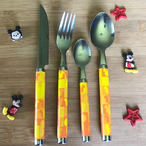 May include: A set of four yellow and orange Mickey Mouse themed cutlery pieces, including a knife, fork, and two spoons. The handles feature a repeating pattern of Mickey Mouse heads.
