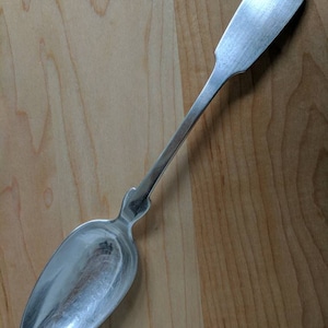 May include: A silver spoon with a rounded bowl and a long, slender handle. The spoon is lying on a wooden surface.