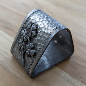 May include: A silver napkin ring with a floral design. The ring is textured and has a hammered finish.