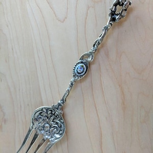 May include: Silver-toned fork with an ornate design and a small enamel medallion featuring a floral motif. The handle is topped with a stylized animal figure.