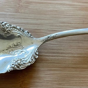 Może przedstawiać: Srebrna łyżka do serwowania z ozdobnym wzorem kwiatowym. Łyżka ma szeroką, zaokrągloną miskę i długi, smukły trzonek.