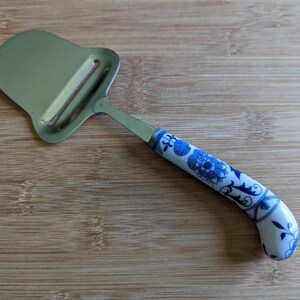 Puede incluir: Un rallador de queso de acero inoxidable con un mango de cerámica blanca decorado con un patrón floral azul.