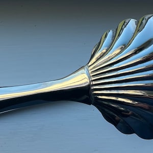 May include: Silver-toned serving tongs with a shell-shaped head. The tongs are designed to resemble a scallop shell.
