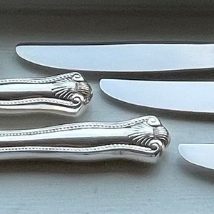 May include: Five silver butter knives with ornate handles. The knives have a polished finish and are arranged in a row.