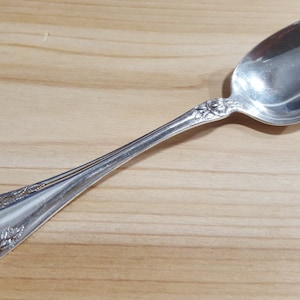 May include: A silver spoon with a floral design on the handle and bowl. The spoon is resting on a light-colored wooden surface. The spoon's bowl is oval-shaped and reflects the surrounding light.
