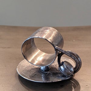 May include: Silver napkin ring shaped like a teacup with a saucer. The cup has a floral design and the saucer has a leaf design.