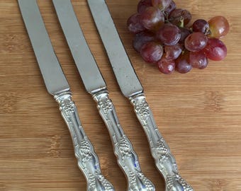 May include: Three silver butter knives with ornate handles, arranged on a wooden surface next to a bunch of red grapes. The knives have a decorative grape and leaf design on the handles.