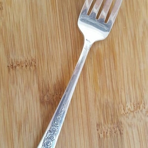Op de afbeelding: Een zilveren vork met een sierlijk bloemmotief op het handvat. De vork ligt op een houten oppervlak.