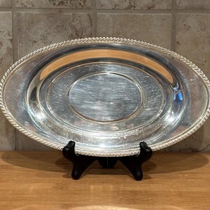 Può includere: Vassoio ovale d'argento da portata con bordo a corda. Il vassoio è esposto su un supporto nero, evidenziando la sua superficie riflettente. Il design è semplice ed elegante, adatto per servire cibo o come elemento decorativo.