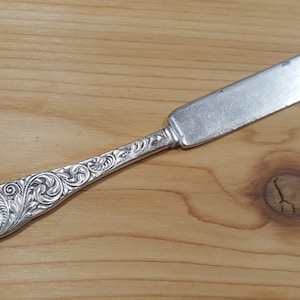 May include: A silver butter knife with an ornate, floral design on the handle. The blade is rectangular and the handle features a detailed, swirling pattern. The knife is set against a light brown wooden surface.