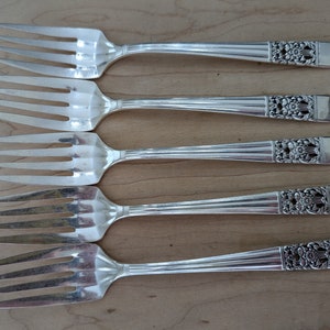 May include: Five silver forks with ornate handles. The forks are arranged in a row, with the handles pointing to the right. The forks are all the same size and shape.