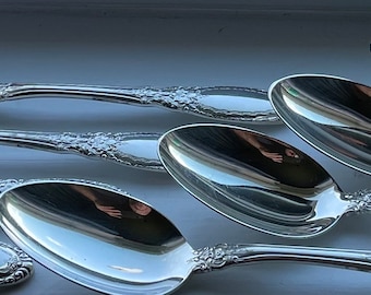 May include: A set of eight silver spoons with ornate handles. The spoons are arranged in a staggered pattern, with the handles overlapping.
