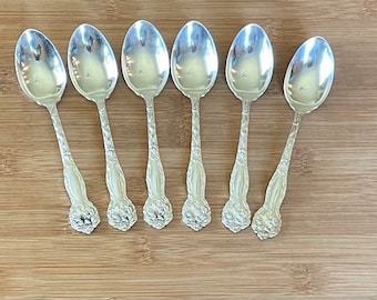 May include: Six silver spoons with ornate handles. The spoons have a floral design and are arranged in a row on a wooden surface.