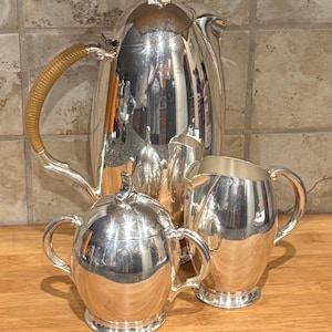 May include: A silver tea set with a teapot, creamer, sugar bowl, and a small pitcher. The teapot has a woven handle. The set is displayed on a wooden surface against a tiled background. The silver reflects the surrounding environment.