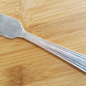 May include: A silver butter knife with a decorative pattern on the handle. The knife is lying on a wooden surface.