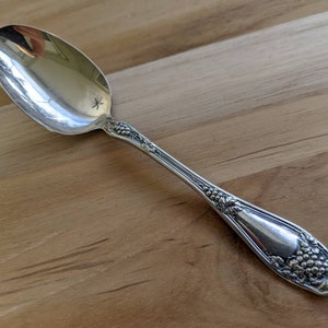 May include: A silver spoon with an ornate design featuring grapes and leaves. The spoon has a polished finish and a slightly tarnished handle.
