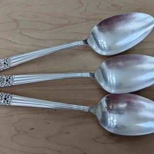 May include: Three silver spoons with ornate handles. The spoons are arranged in a row, with the handles pointing to the left. The spoons are polished and shiny.
