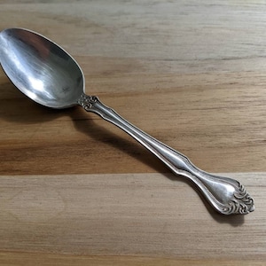 May include: A silver teaspoon with an ornate handle. The spoon is lying on a wooden surface.