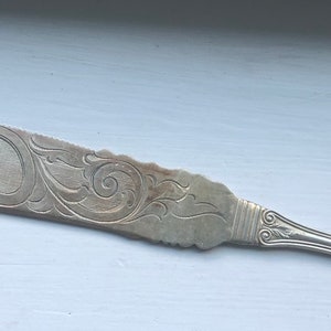May include: A silver butter knife with an ornate, engraved design. The handle is rounded and has a floral pattern.