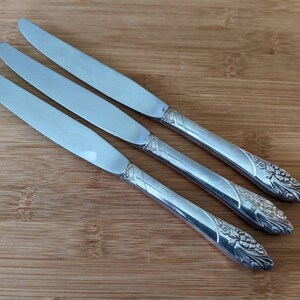 Puede incluir: Tres cuchillos de cena de plata con mangos ornamentados. Los mangos tienen un diseño floral.