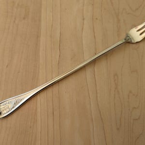 May include: A silver fork with three tines, resting on a light brown wooden surface. The handle has an ornate design at the end. The fork is a piece of silverware.