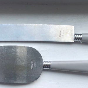 May include: Two silver cake knives with grey handles. The top knife has a serrated edge and the bottom knife has a smooth edge. Both knives have "STAINLESS STEEL" printed on the blade.