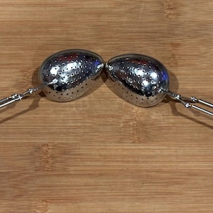 May include: Two stainless steel tea infusers, each with a perforated oval-shaped basket and a long handle. The handles are connected to the basket with a hinge. The infusers are resting on a wooden surface.
