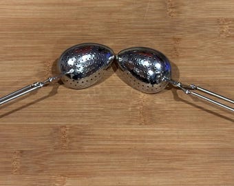 May include: Two stainless steel tea infusers, each with a perforated oval-shaped basket and a long handle. The handles are connected to the basket with a hinge. The infusers are resting on a wooden surface.