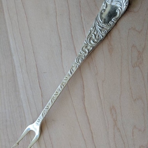 May include: A silver-plated fork with an ornate handle and two prongs. The handle is decorated with a floral design.