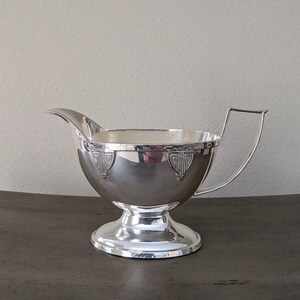 May include: A silver creamer with a hammered finish and a decorative design around the rim. The creamer has a round body and a pedestal base.
